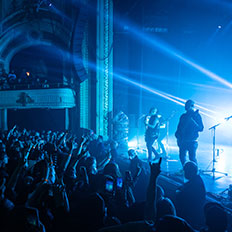 Excited crowd at the Burton Cummings Theatre watching a live performance, with blue stage lights illuminating both the band and the ornate architecture of the venue. Audience members are raising their hands and capturing the moment on their phones.