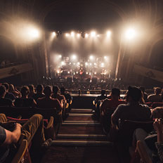View from the upper seats at the Burton Cummings Theatre, looking down at a concert stage with warm, glowing lights. The audience is seated, watching the band perform, while the theatre's elegant, curved balconies frame the scene.