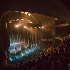 Audience seated at the Burton Cummings Theatre, watching a live performance with warm, atmospheric lighting illuminating the stage and the theatre’s iconic arched ceiling and balconies. The artist performs to a captivated crowd in the historic venue.