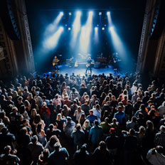 Overhead view of a crowd gathered at the Burton Cummings Theatre for a live concert, with the band performing on a stage illuminated by blue and white spotlights. The theatre’s detailed balconies frame the scene, adding to the intimate concert atmosphere.