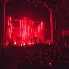 Live performance at the Burton Cummings Theatre, with the stage bathed in vibrant red lighting and vertical light bars creating a dramatic backdrop. The silhouettes of the band are visible as they perform to a packed crowd, with the theatre's ornate architecture surrounding the scene.