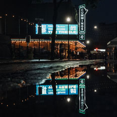 Exterior night view of the Burton Cummings Theatre with its neon sign reflecting in a puddle on the street. The marquee lights and string lights add warmth to the scene, while the event signage advertises upcoming performances.
