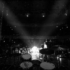 Black-and-white view from behind a drum kit on stage at the Burton Cummings Theatre, showing a performer under bright stage lights with the audience filling the balcony and floor seats. The grand architecture of the venue is subtly visible in the background.