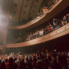 Packed audience at the Burton Cummings Theatre, with fans in both the balcony and main floor sections cheering and raising their hands during a live concert. The venue's grand architecture is illuminated by soft lighting, highlighting the arched ceiling and intricate balcony details, creating an electric atmosphere.