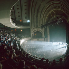 Wide-angle view of the interior of the historic Burton Cummings Theatre, filled with a seated audience watching a live performance. The ornate architecture, with its arched ceiling and detailed balconies, frames the stage, where beams of light cut through the atmosphere, creating a dramatic scene.