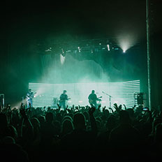Concert stage with a band performing in front of a packed crowd, illuminated by green and white horizontal lights in the background, creating a vibrant atmosphere. The audience raises their hands, enjoying the live music in a dark, atmospheric venue.